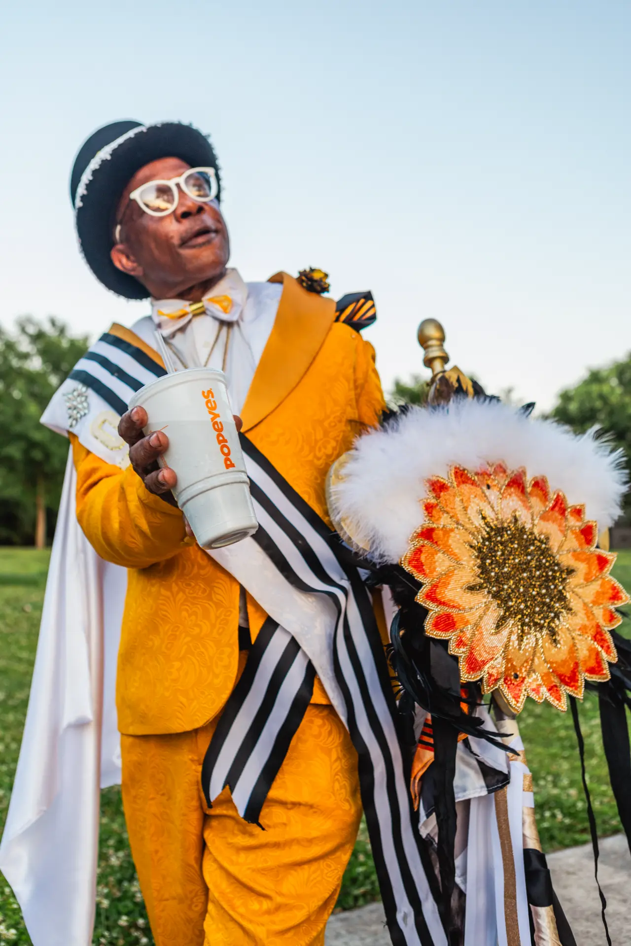A Mardi Gras grand marshal in a golden suit and white cape holding a Popeyes cup