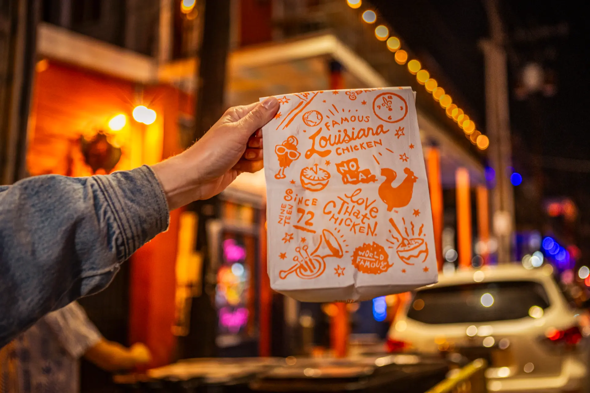 A hand holding a Popeyes Famous Louisiana Chicken paper bag against festive night lights