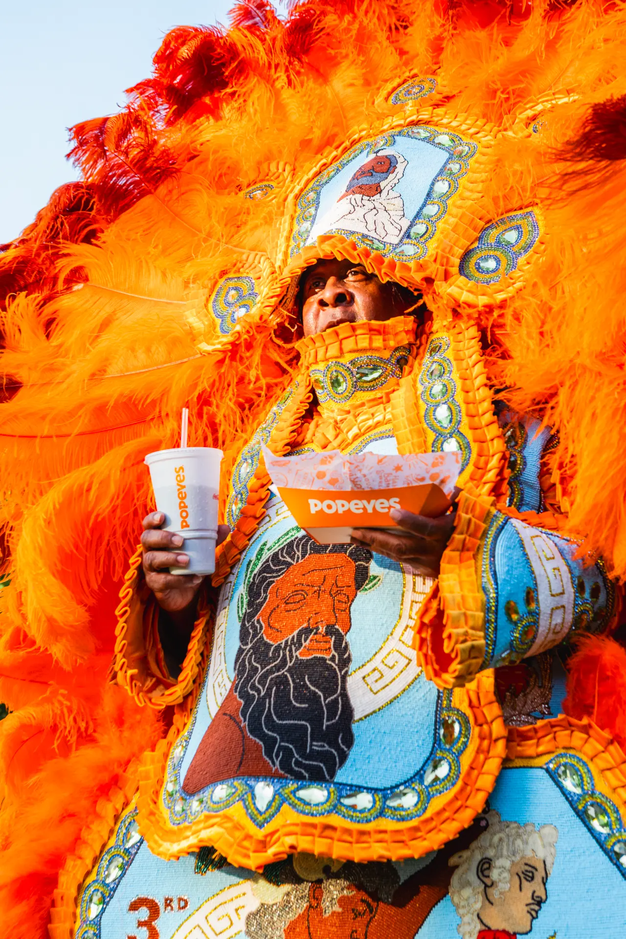 A Mardi Gras performer in vibrant orange feathered regalia holding a Popeyes drink and box