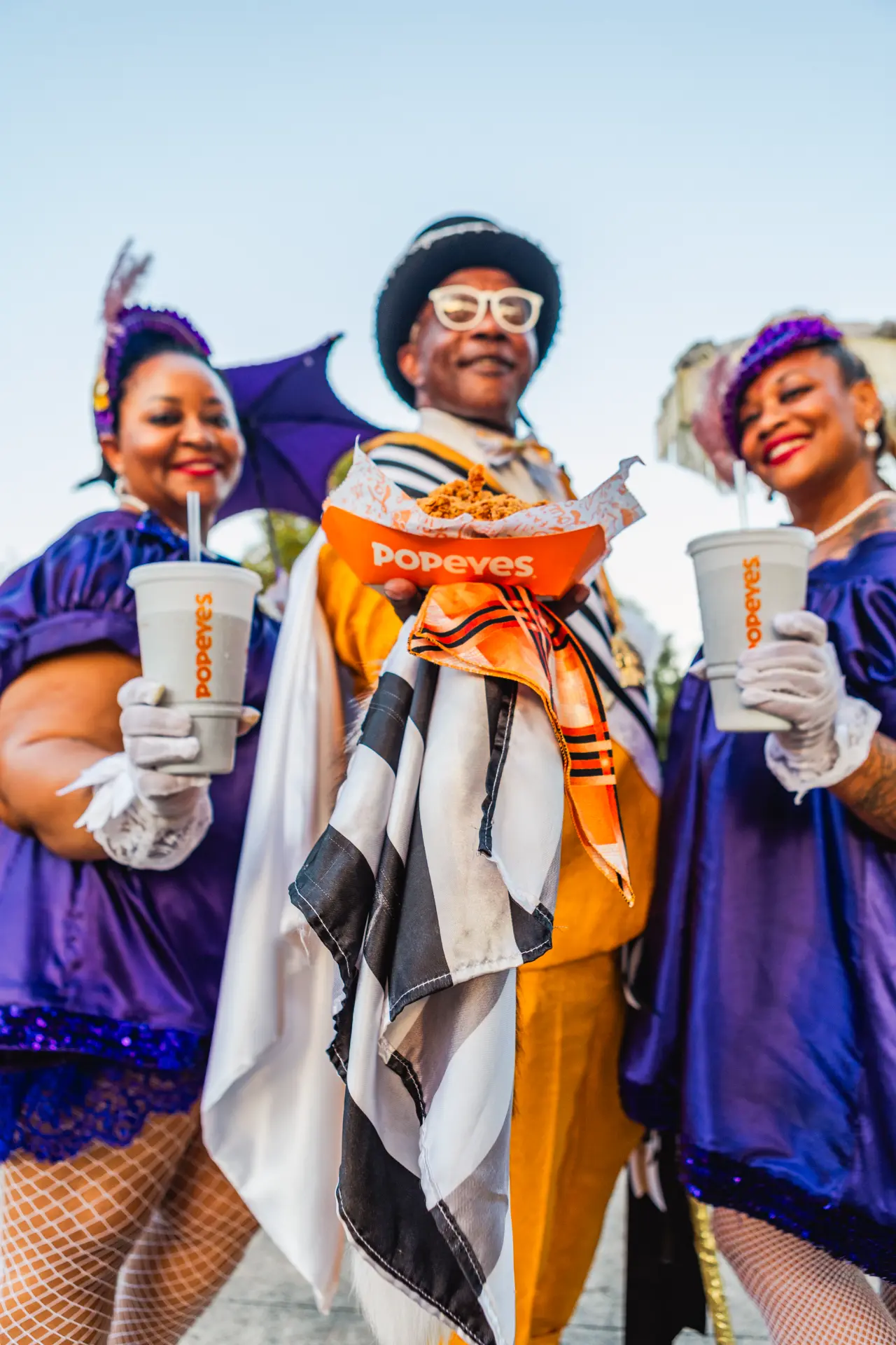 Three Mardi Gras revellers in costume holding Popeyes drinks and chicken