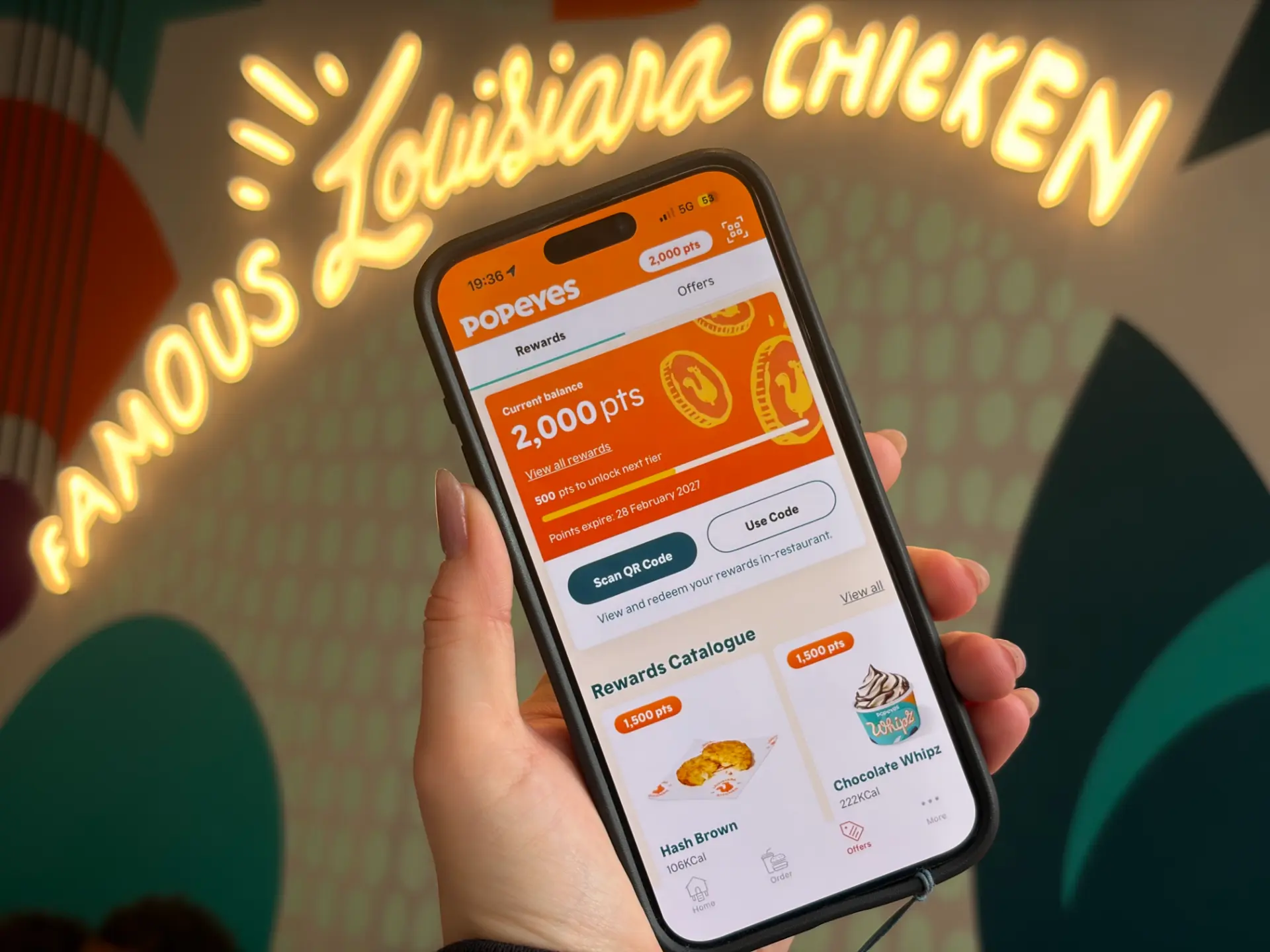 A hand holding a phone showing the Popeyes Rewards app in front of a glowing Famous Louisiana Chicken sign